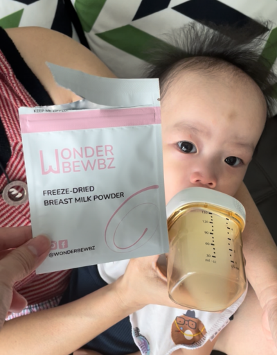 Person holding a package of Wonder Bewbz freeze-dried breast milk powder with a baby drinking from a bottle in the background.
